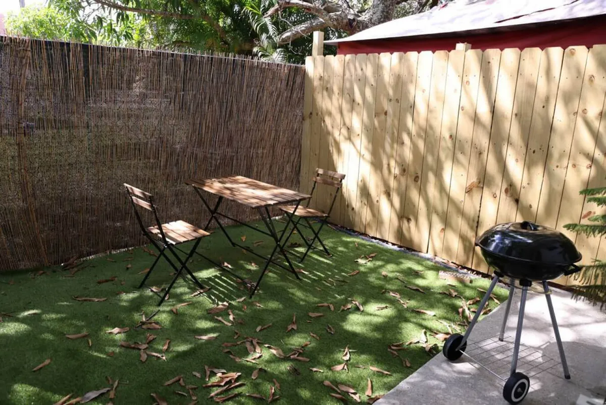 An inviting outdoor patio scene with a wooden table and black grill set against a backdrop of a bamboo fence and green grass. The setup suggests possibilities for grilling, outdoor dining, and enjoying meals in a relaxed setting, reflecting the option to dine al fresco just outside the kitchen.