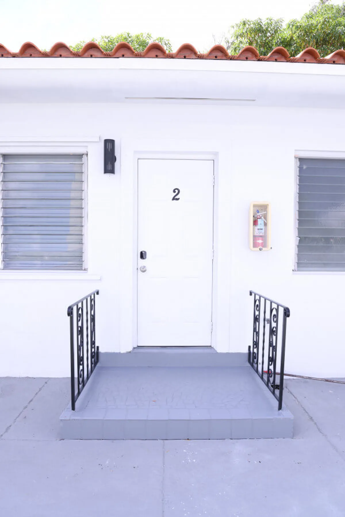 The image showcases the exterior of a ground-floor Miami apartment, featuring a private white door with the number 2, flanked by two windows with horizontal blinds. A small, tiled porch with elegant black wrought iron railings offers a charming, welcoming entryway. The porch area suggests a serene and well-kept space, perfect for relaxing before stepping inside. A fire extinguisher mounted on the wall highlights attention to safety.