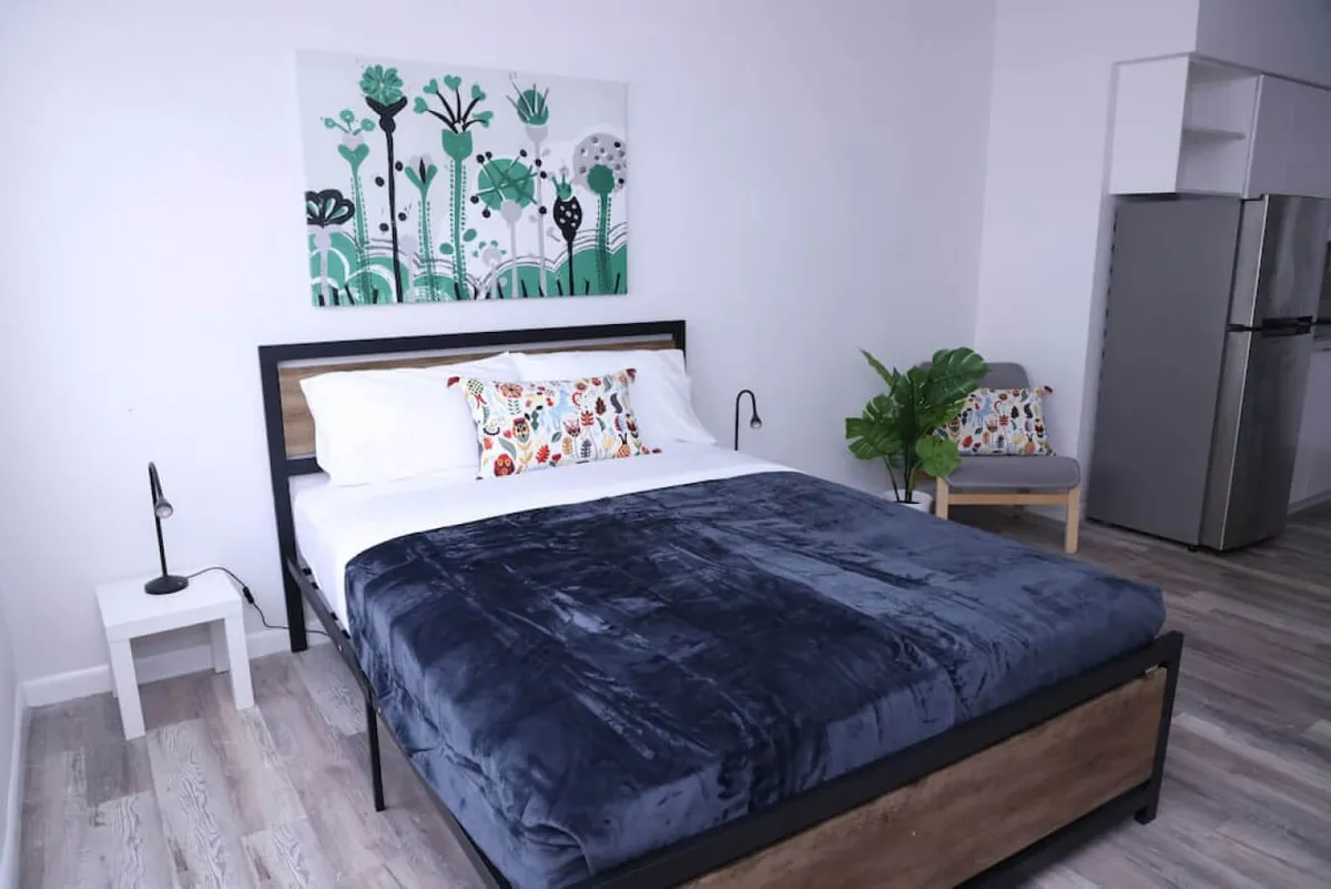 A serene studio interior featuring a large bed adorned with a dark blue blanket and decorative pillows, modern botanical artwork above the bed, a compact kitchen area with stainless steel refrigerator, and a comfortable chair with a colorful cushion beside a potted plant. The space is bathed in soft natural light, combining sleeping, lounging, and kitchen amenities in a single open-plan area.