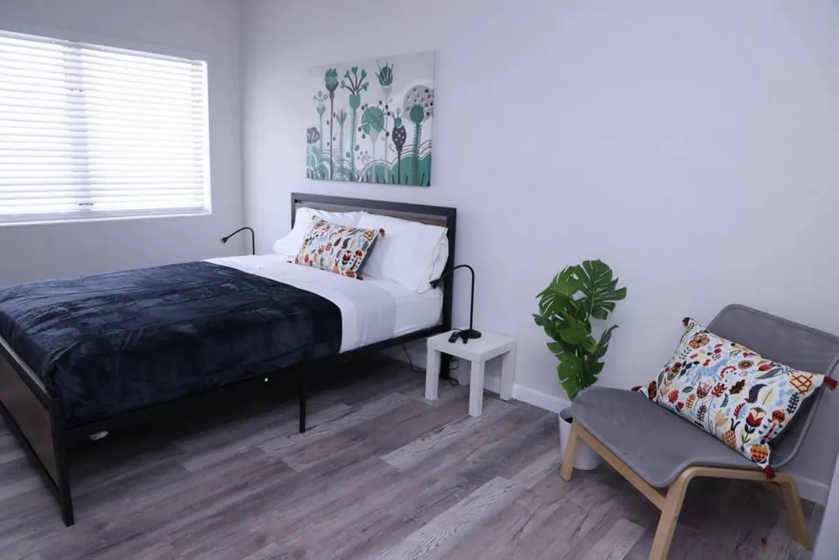A cozy bedroom scene featuring a bed dressed in a dark navy blanket with decorative pillows, a modern chair accented with a colorful pillow, and a green potted plant beside the chair. Sunlight filters through window blinds, creating a warm, inviting atmosphere.