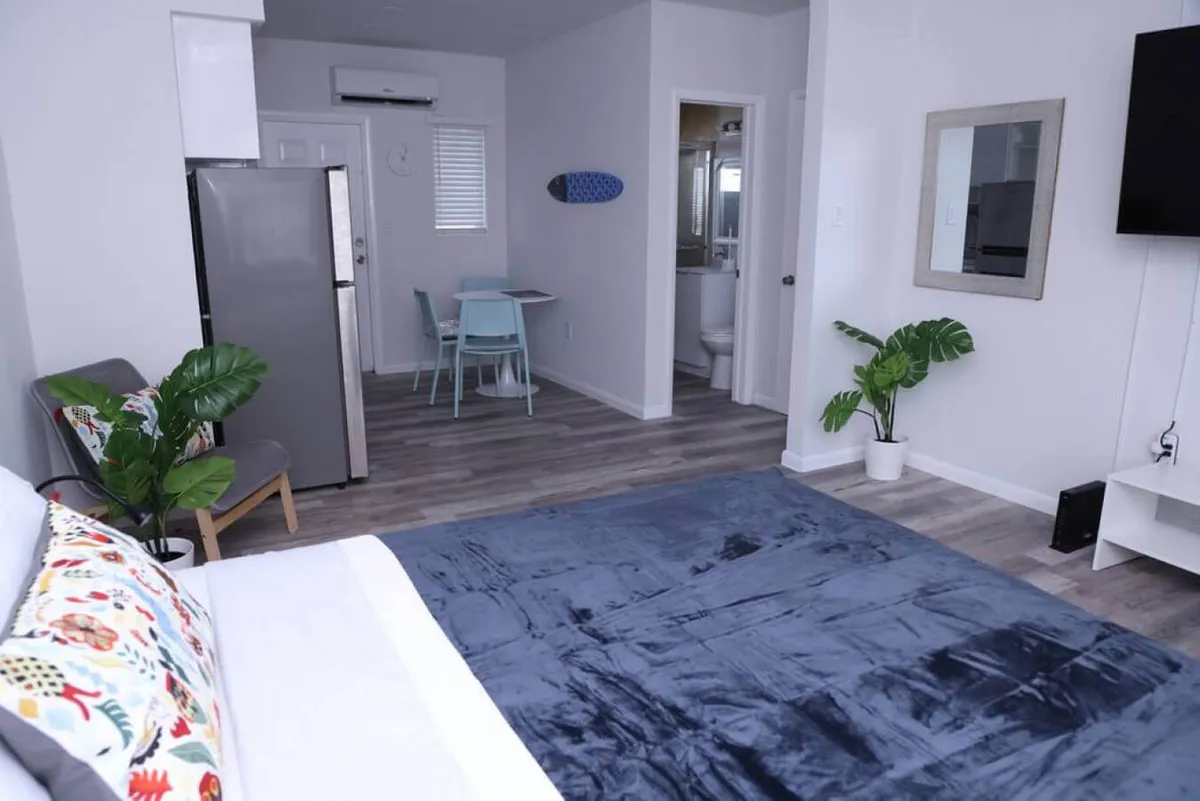 A bright and inviting studio interior with a large bed featuring colorful pillows, a flat-screen smart TV mounted on the wall, a dining table with chairs, and a view toward the bathroom entrance. The space blends areas for rest, entertainment, and dining, all in a modern, thoughtfully arranged environment.