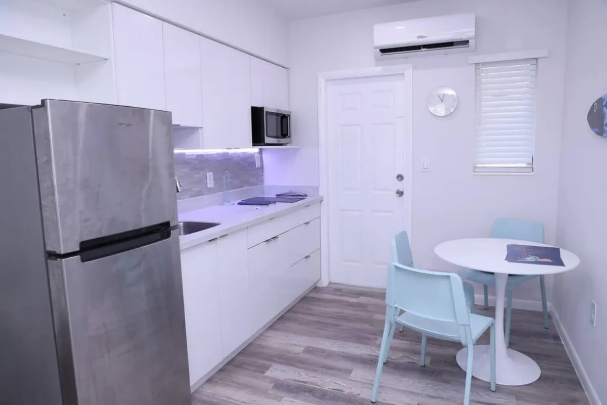 A bright and modern kitchen scene featuring a stainless steel refrigerator, sleek white cabinetry with under-cabinet lighting, and a small round dining table surrounded by pastel blue chairs. The space is finished with a wall-mounted air conditioning unit and opens to a private patio area, making it a welcoming spot for both meal preparation and dining.