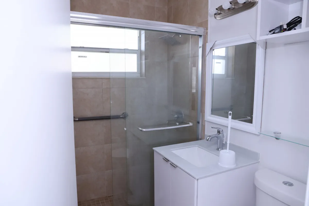 A modern, well-lit bathroom featuring a glass-enclosed shower with tiled walls, a clean white vanity and sink, a large mirror above, and a toilet. The space is fresh and organized, ideal for daily routines.